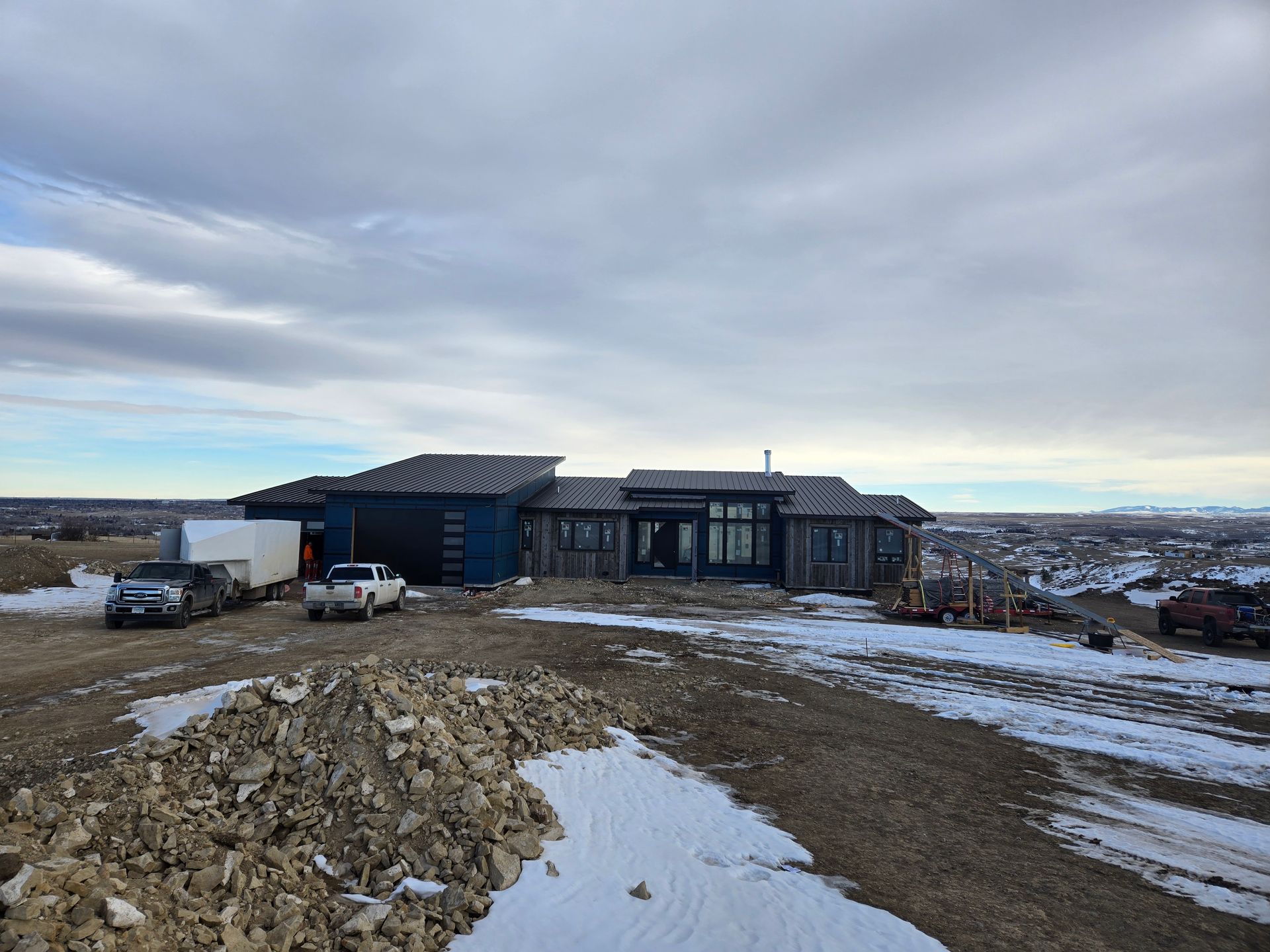 A partially built blue house on a hill, construction equipment and vehicles present, cloudy sky.