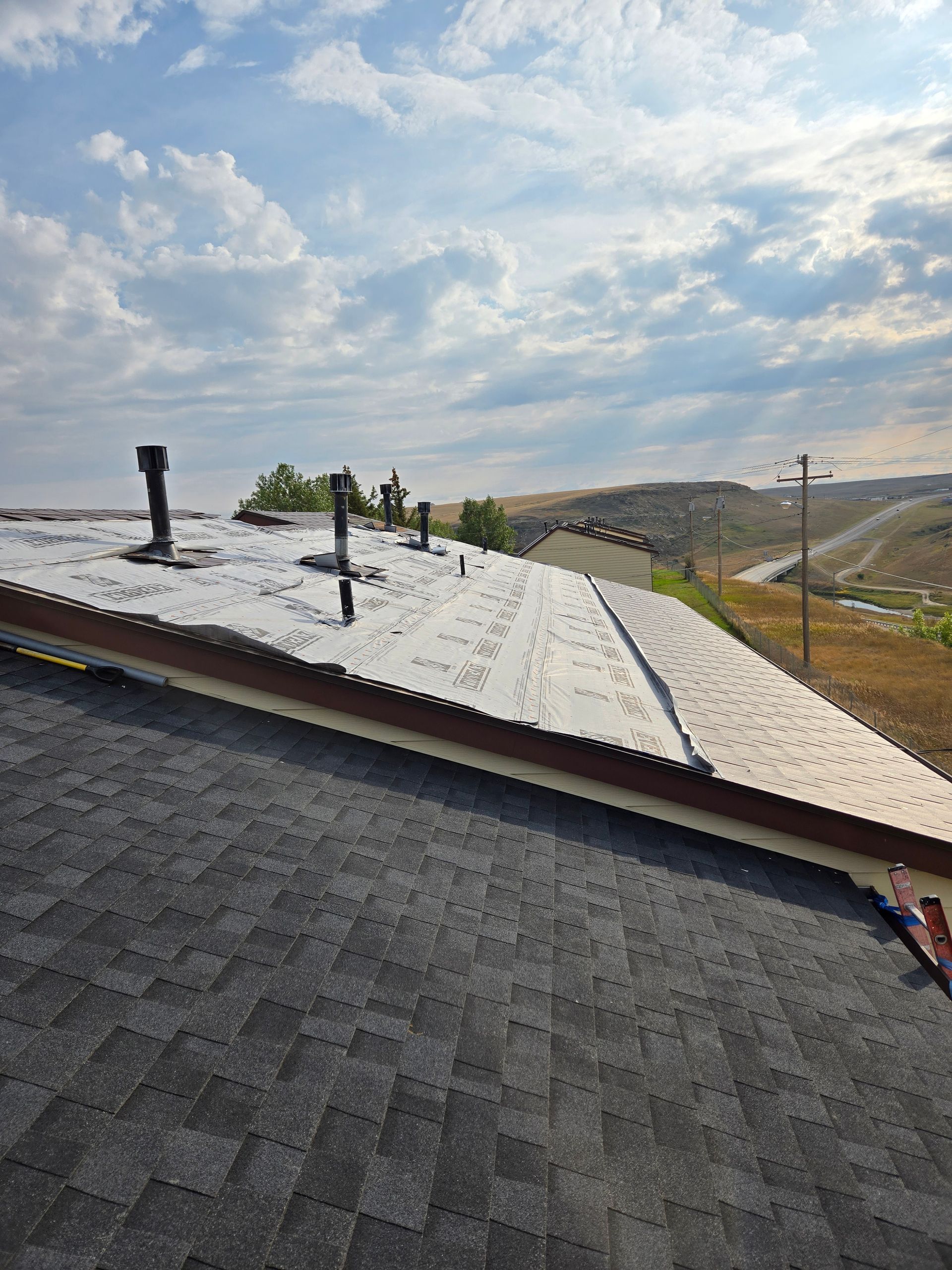 A roof with shingles and a section of flat roofing. Several vents and a brown edge. Cloudy sky.