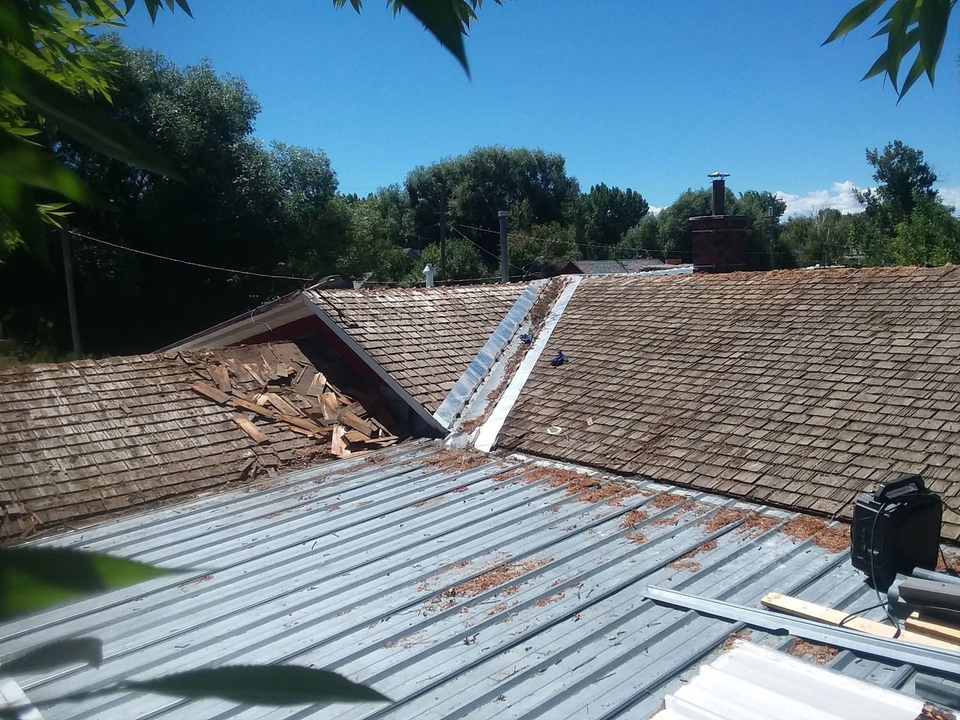 Roof with missing shingles and metal sheeting, surrounded by trees under a blue sky.