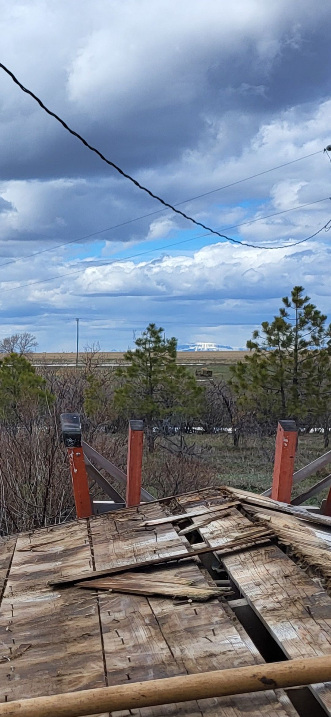 View from a damaged rooftop. Power lines, trees, cloudy sky, and distant landscape are visible.