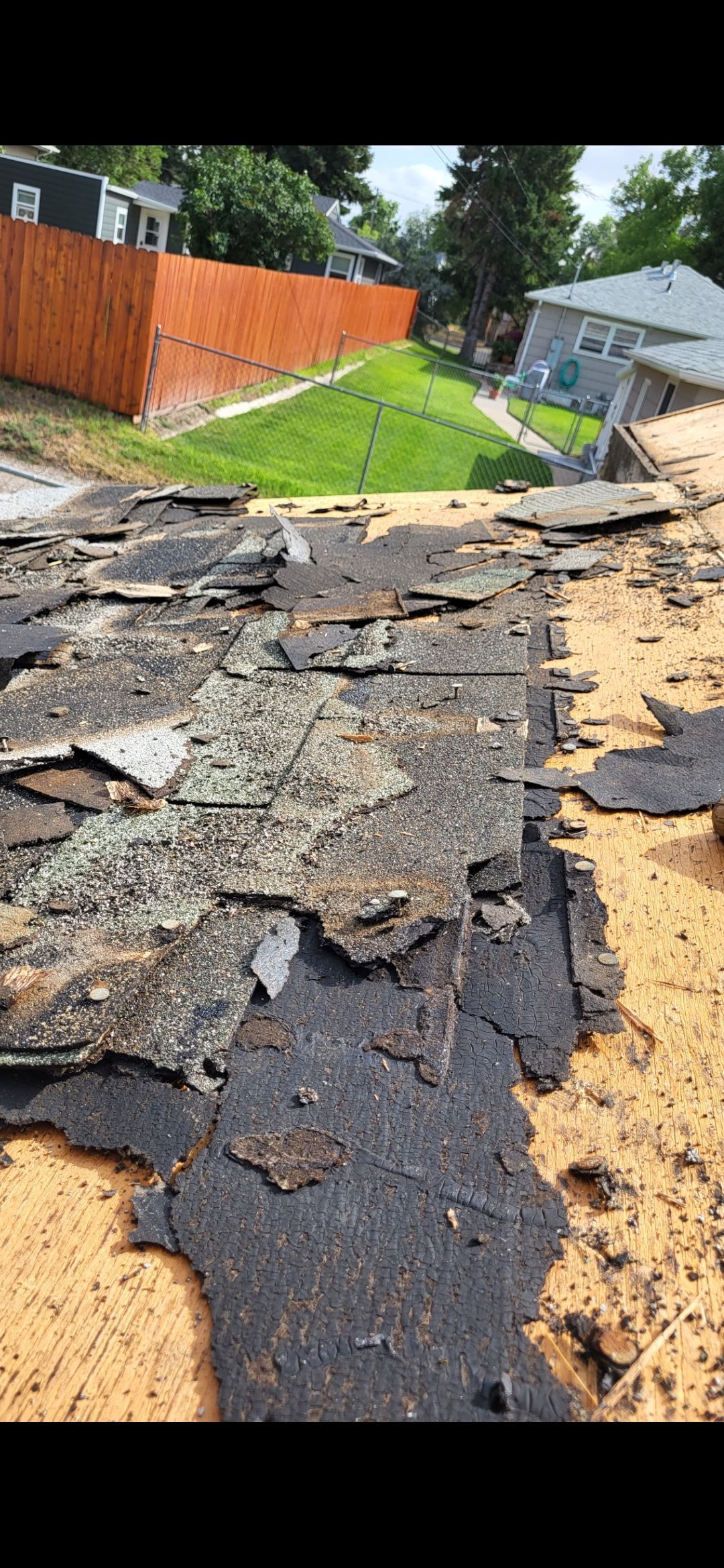 Close-up of a damaged roof with crumbling shingles, showing a yard and houses in the background.