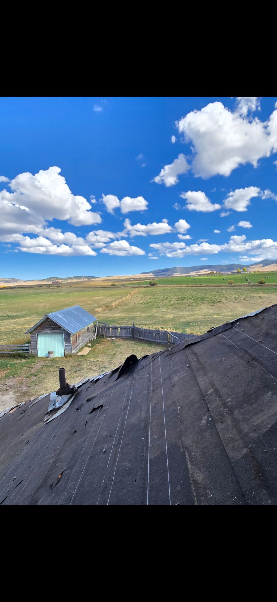 Blue sky with clouds over a rural landscape; a dilapidated building with a metal roof is in the distance.