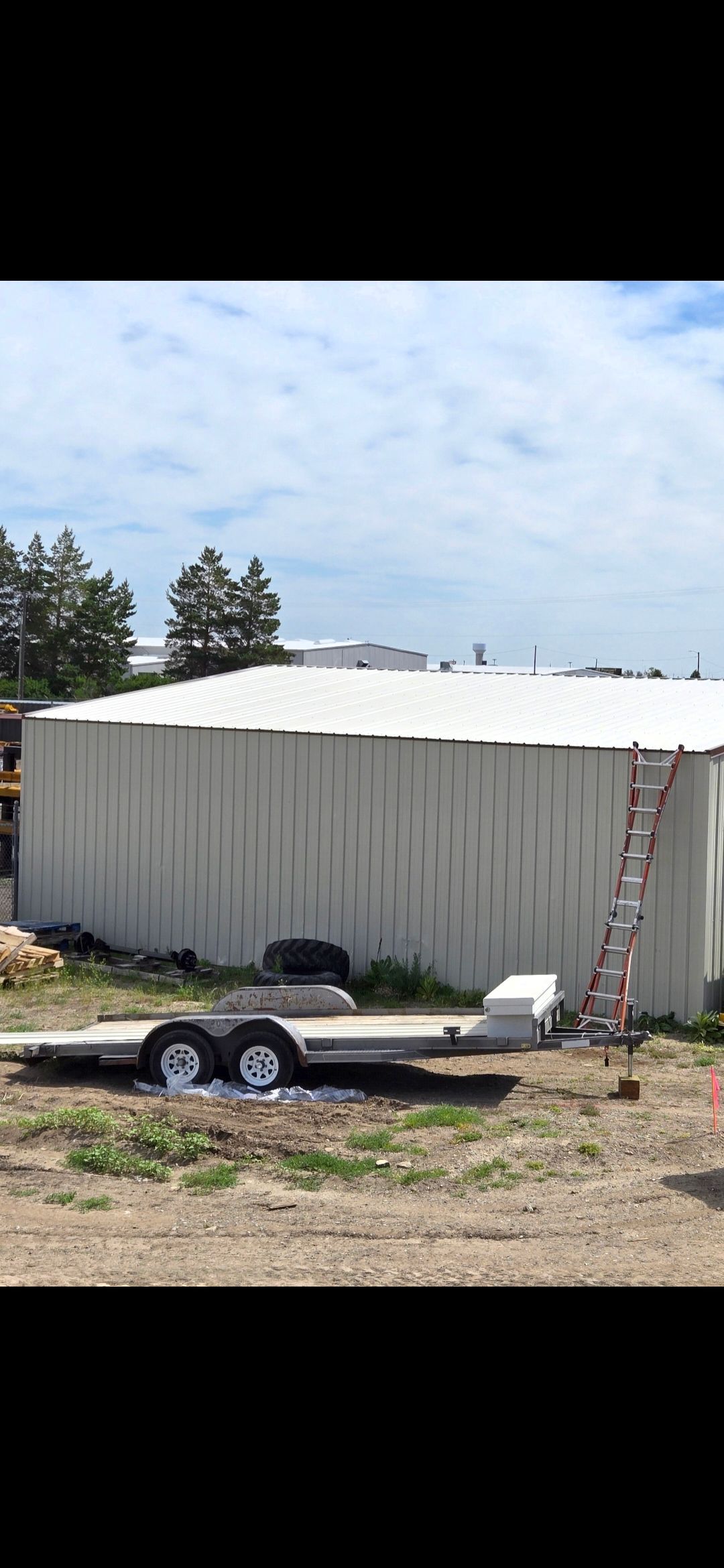 A metal building with a ladder leans against it. A trailer is in front. Trees and a cloudy sky in the background.