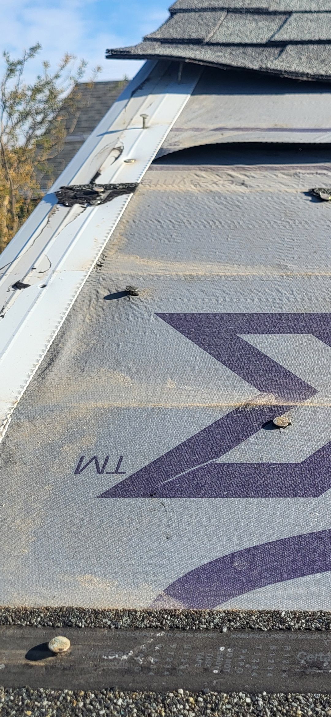 Close-up of a damaged roof with exposed underlayment and purple logo, the word 