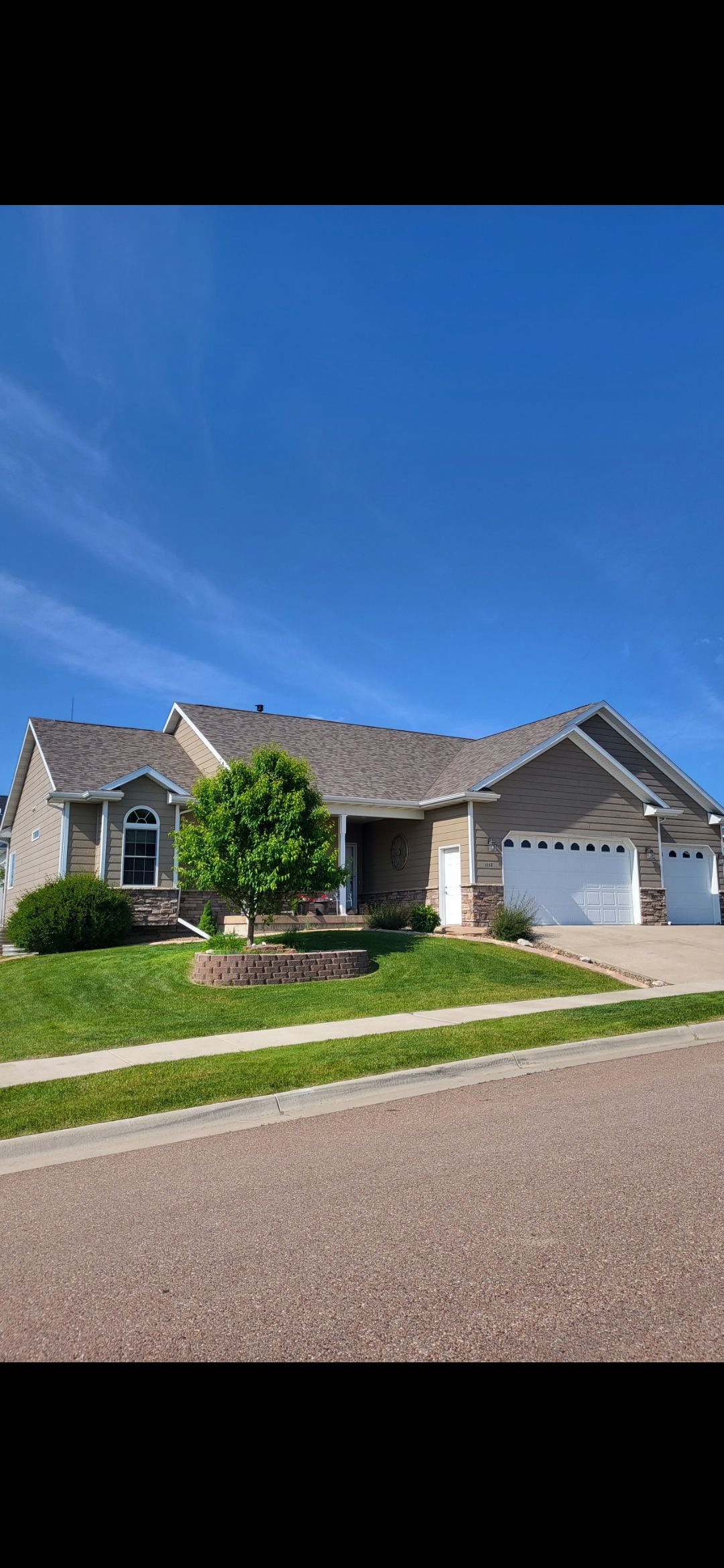 House with a blue sky background. Green lawn in front with a tree and garage doors.