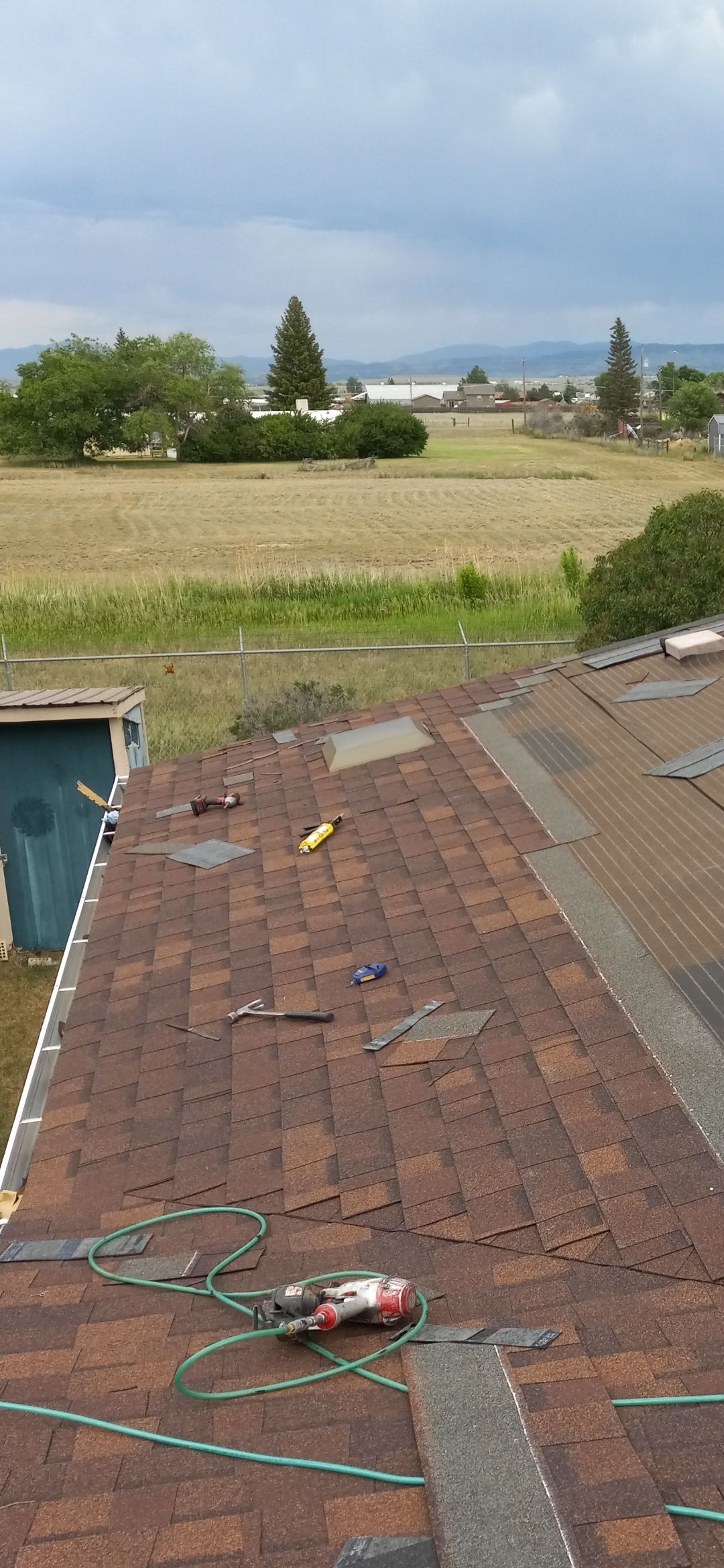 A partially shingled roof with tools on it, overlooking a field and distant buildings under a cloudy sky.