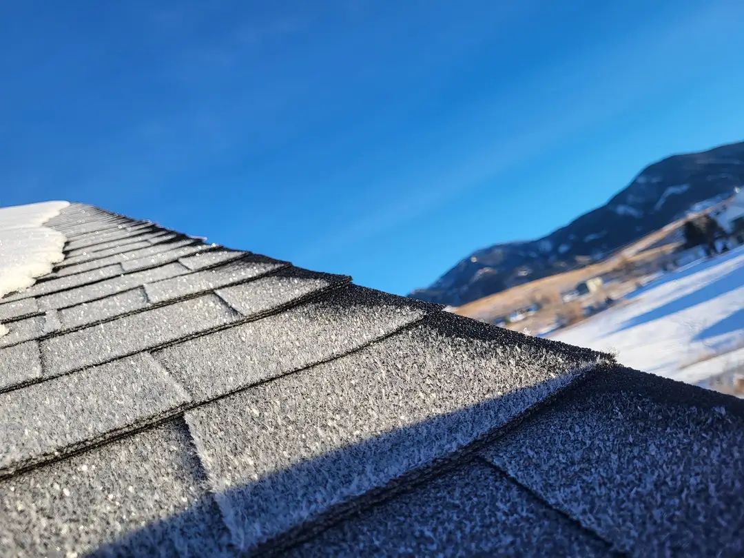 Snowy roof shingles with a mountain backdrop under a blue sky.