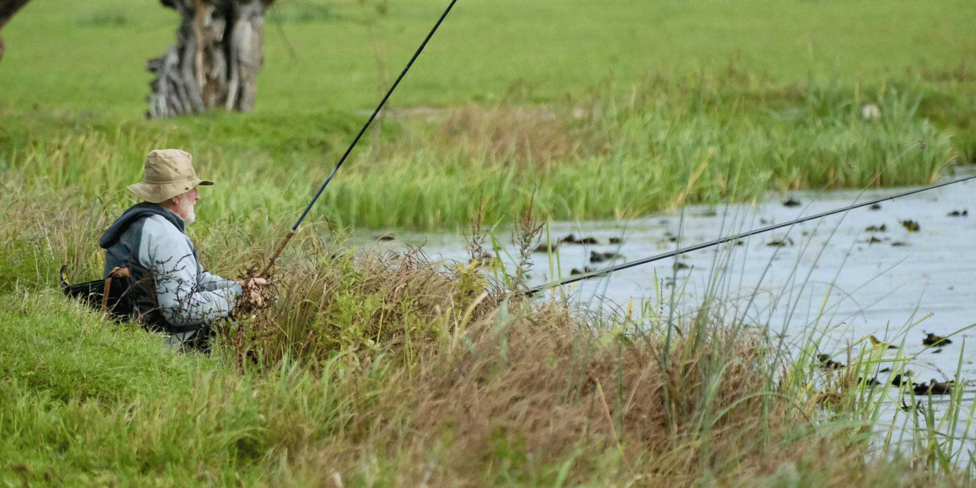 An older man wearing a straw hat, fishing in a grassy area next to a body of water.