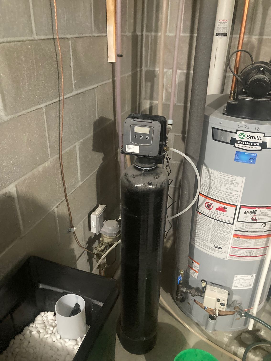 Water softener and hot water heater in a basement setting. Black tank with digital display. Gray cinder block wall.