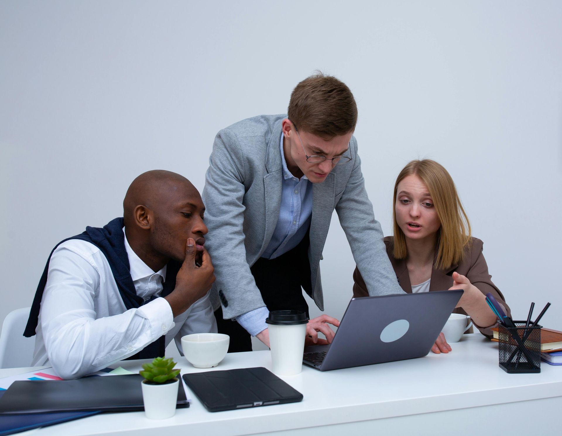 Three people collaborating at a desk, looking at a laptop. One person points, another is thoughtful.