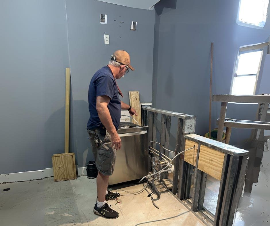 Man in workshop, examining a metal structure with plumbing, wearing a hat and safety glasses. Gray walls, unfinished.
