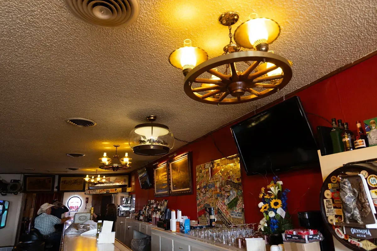 Bar interior with a long counter, red wall, wagon wheel chandelier, and patrons.