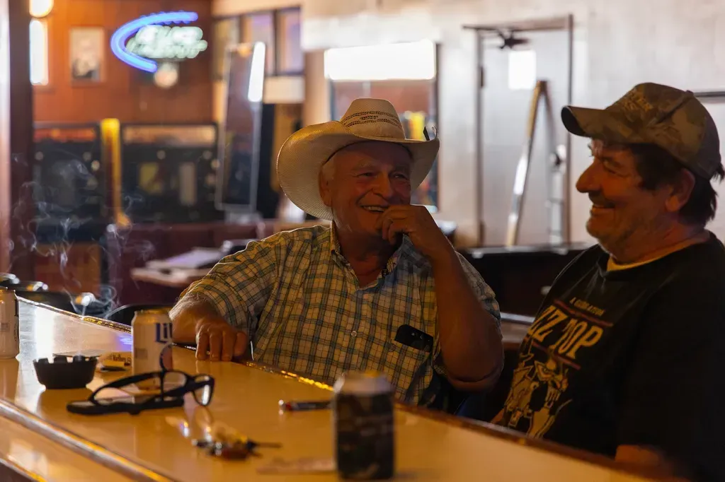 Two men at a bar; one in a cowboy hat smiles, the other laughs. Dim lighting, bar setting.
