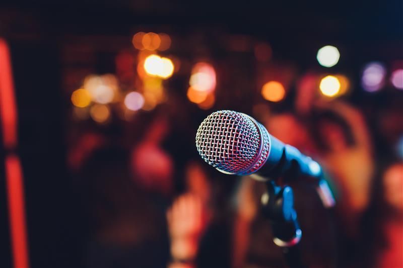 Microphone on stand in front of blurred, lively crowd, bathed in warm lights and dark shadows.