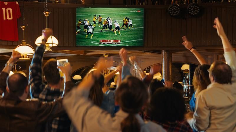 People cheering, watching a football game on a large screen in a bar.