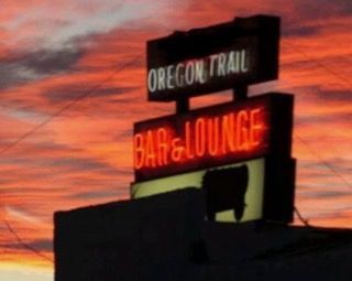 Oregon Trail Bar & Lounge sign at sunset, red neon text, dark building silhouette.