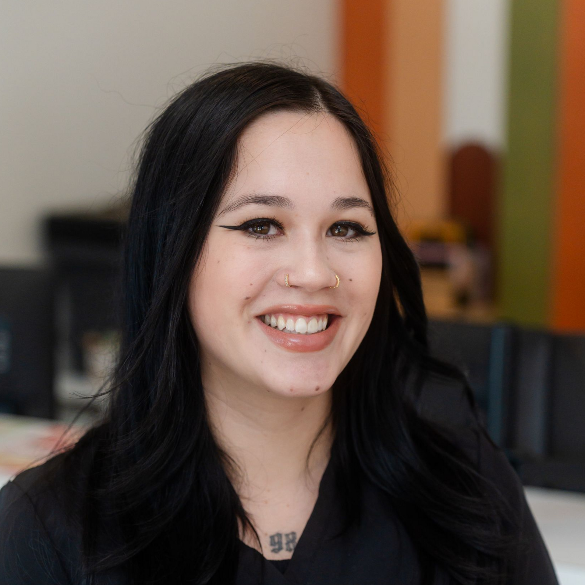 Woman with long black wavy hair smiling at the camera. She's wearing a black top. Background includes an office.