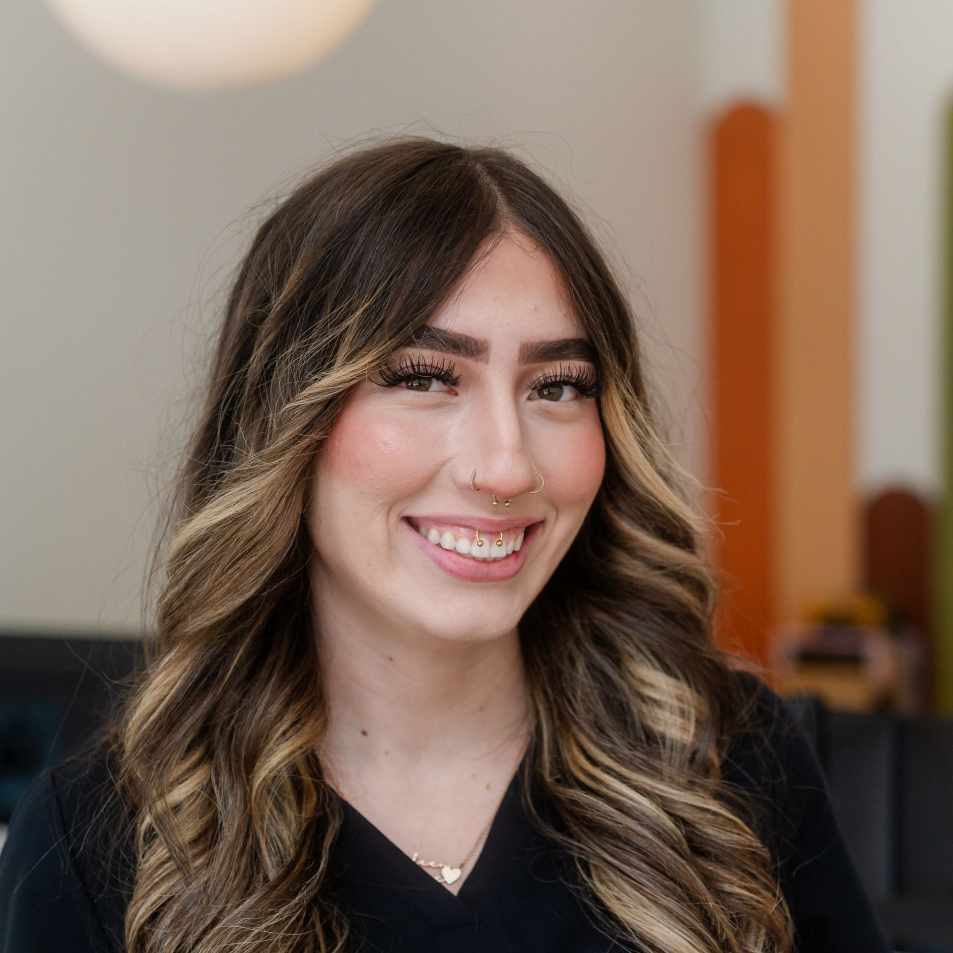 Smiling woman with long, wavy brown hair, nose ring, wearing a black shirt, indoors, soft lighting.