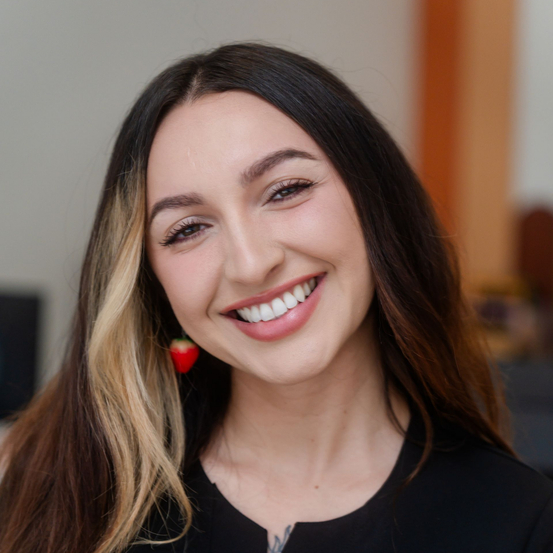 Woman smiling at the camera with long brown hair, a blonde streak, and a strawberry earring.