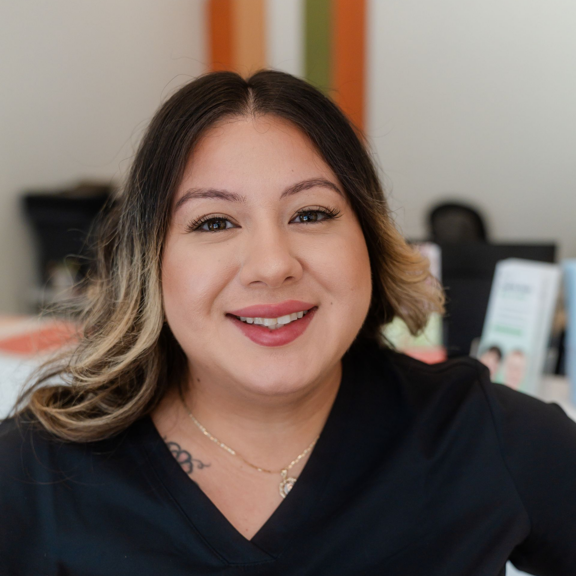 Woman smiling, with dark hair, light highlights, and wearing a black top. Background with blurred office setting.