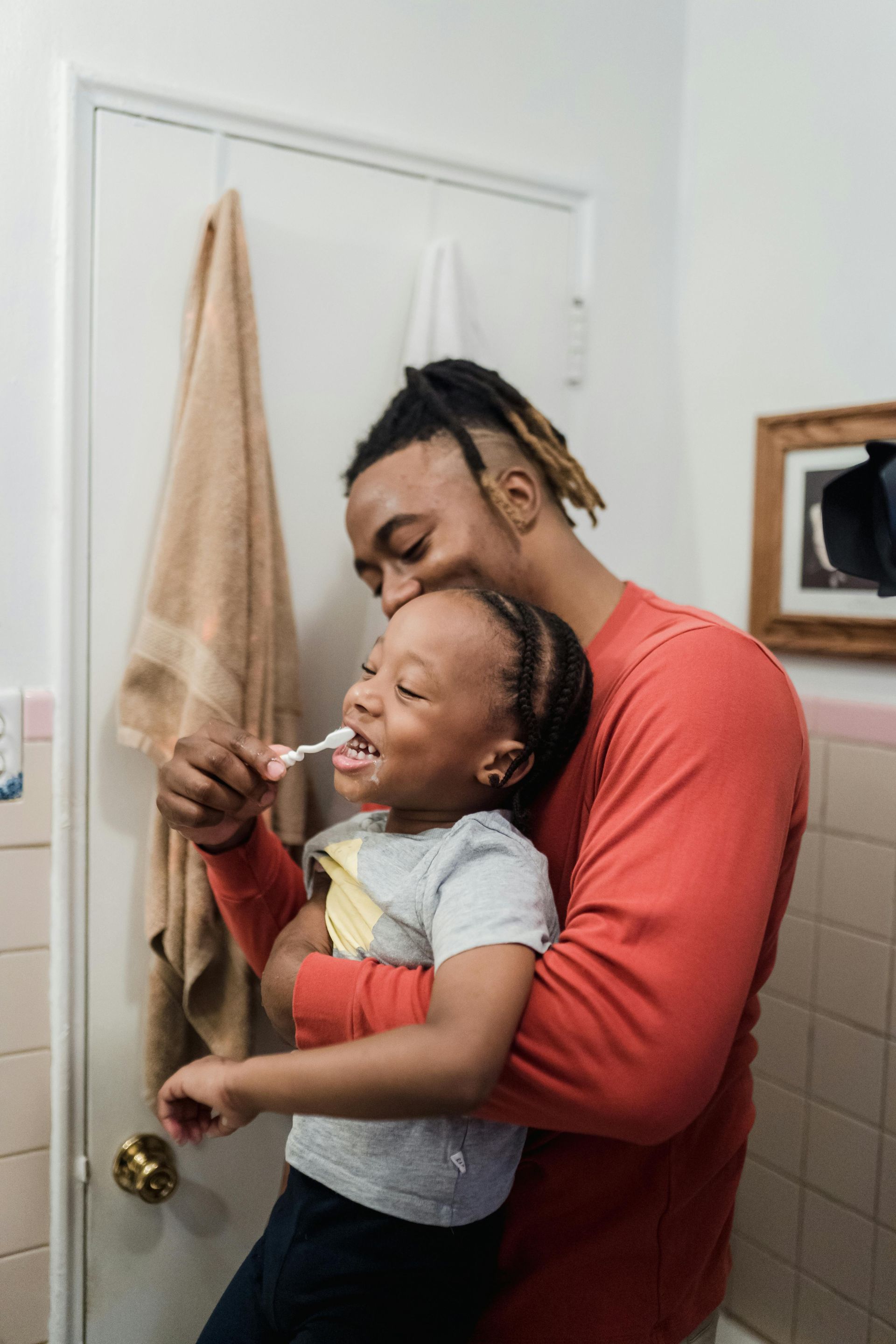 Man holding child, brushing teeth in bathroom. Door and towel visible.