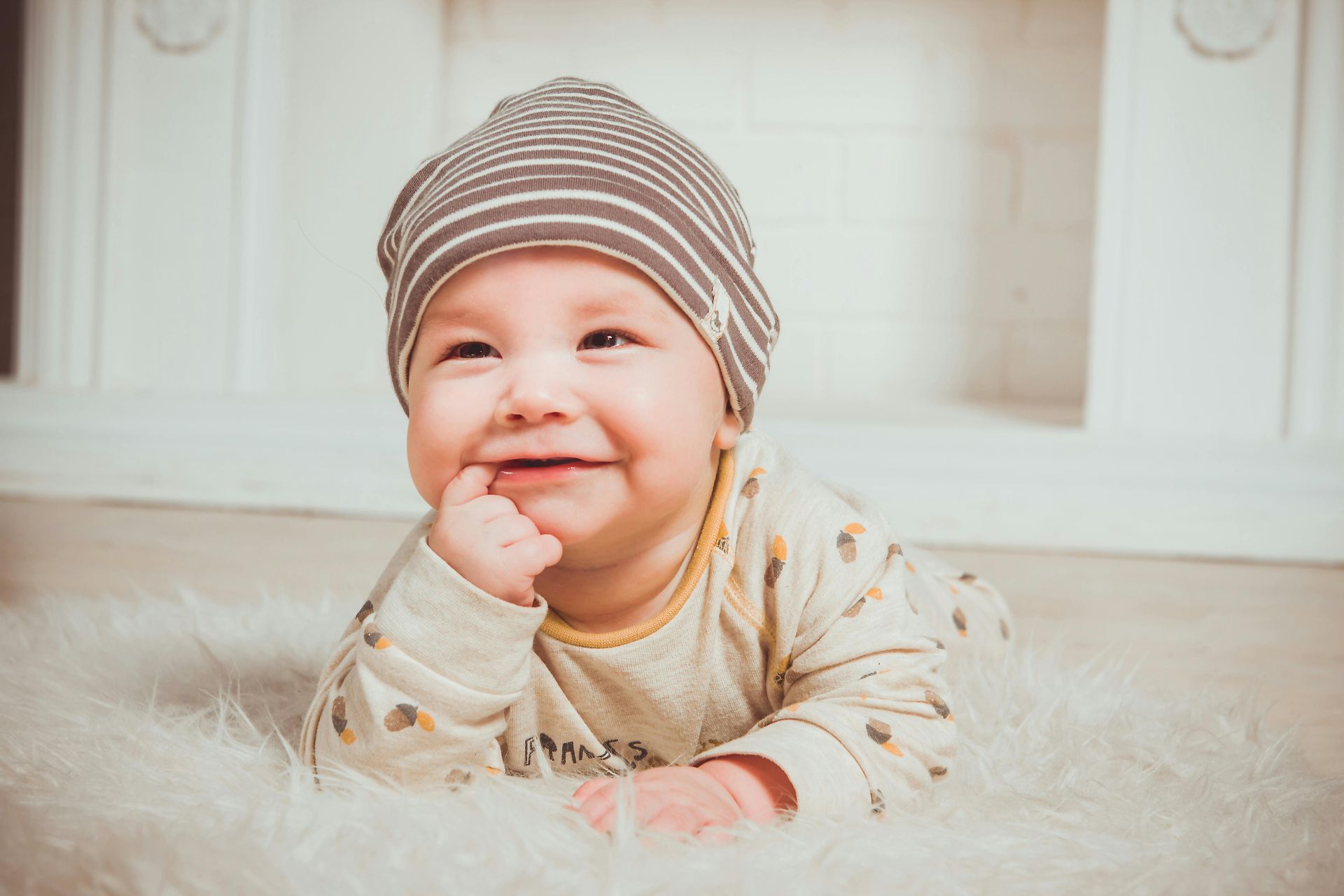 Smiling baby wearing a striped hat and onesie, lying on a white fluffy rug.