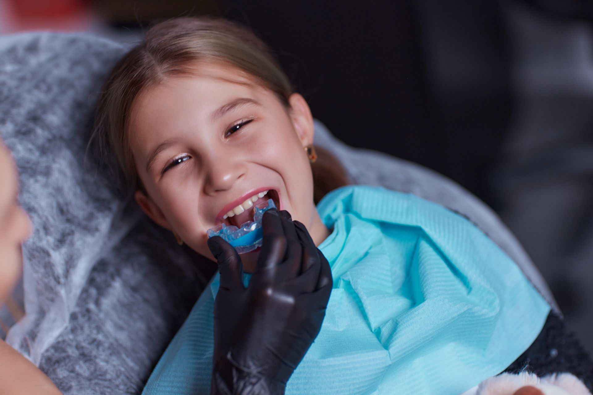 Girl at dentist, wearing blue bib, holding a blue dental impression tray in mouth, with gloved hand assisting.