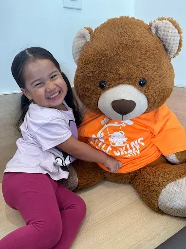 A smiling child hugs a large brown teddy bear wearing an orange shirt; indoors on a mat.
