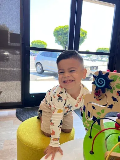 Smiling child on a yellow stool near a window with toys.