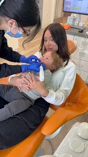 Dentist examining a child's teeth while held by a person in a dental chair.