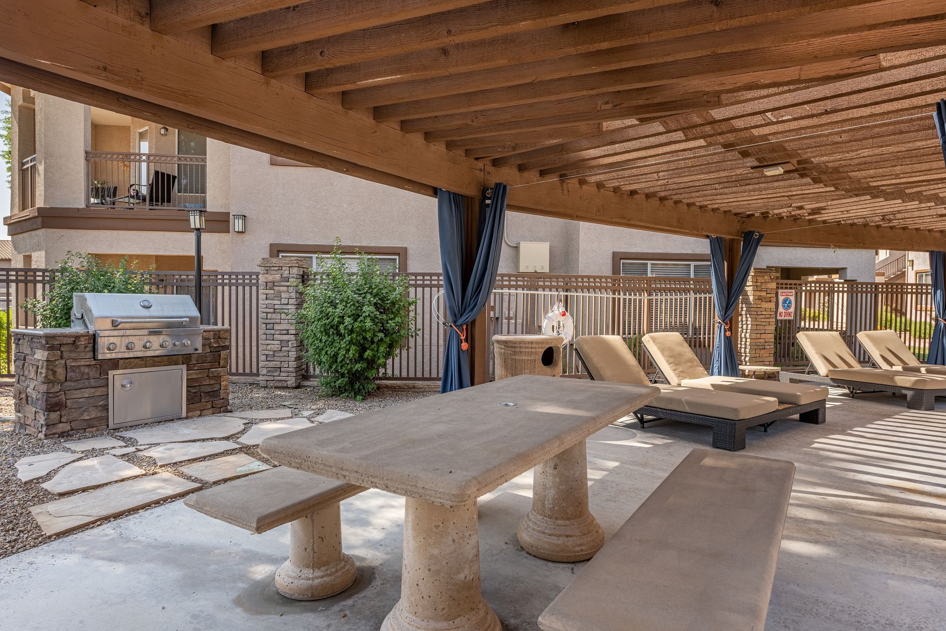 Covered outdoor patio with stone table, benches, lounge chairs, and a grill under a wooden roof.