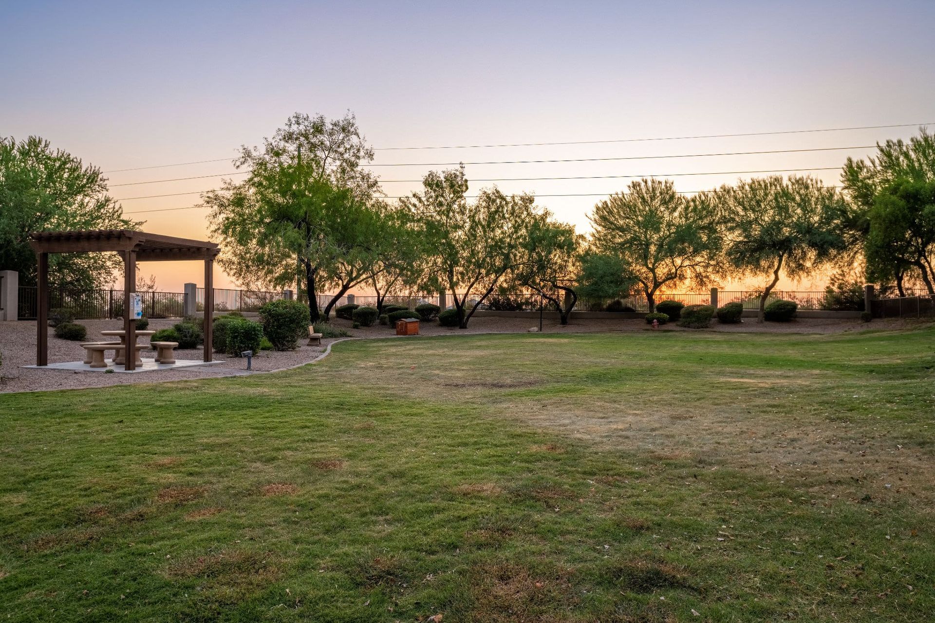 Tranquil park scene at sunset with the sky showing pastel hues of pink and blue. In the foreground, a well-maintained grass lawn leads to a small pavilion with a picnic table. Mature trees dot the landscape, and beyond the foliage, the sun dips below a distant horizon, partly obscured by a tall fence. No people are visible, adding to the peaceful ambiance of the setting at Azure Creek in North Phoenix, AZ.