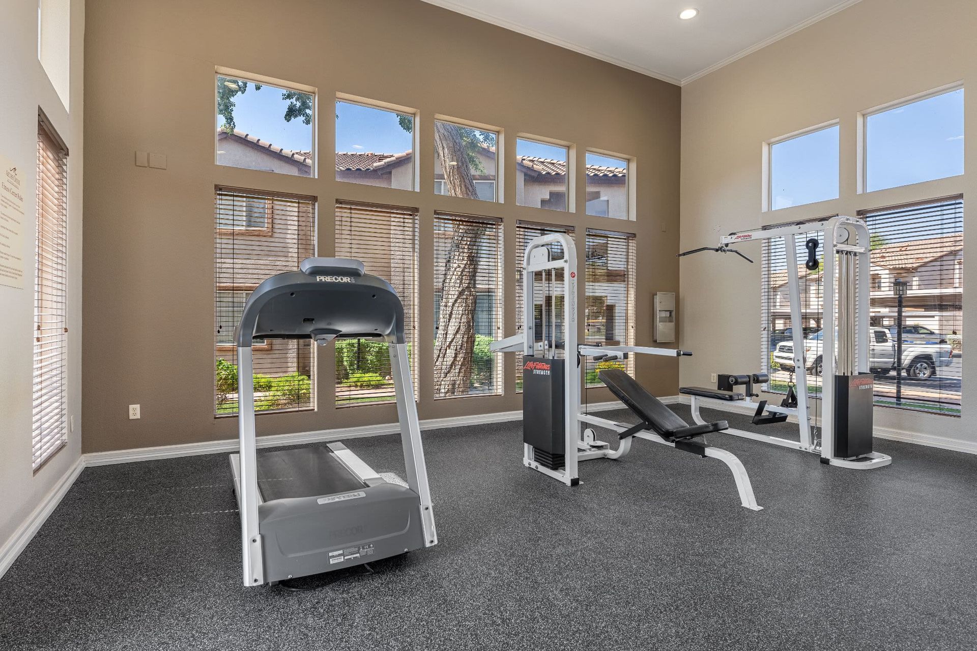 Interior of a modern home gym with high ceilings and tall windows with blinds. The room features a treadmill, a weight bench, and a multi-exercise cable machine, with a view of residential buildings outside the windows. The gym has a dark speckled floor and tan walls at Azure Creek in North Phoenix, AZ.