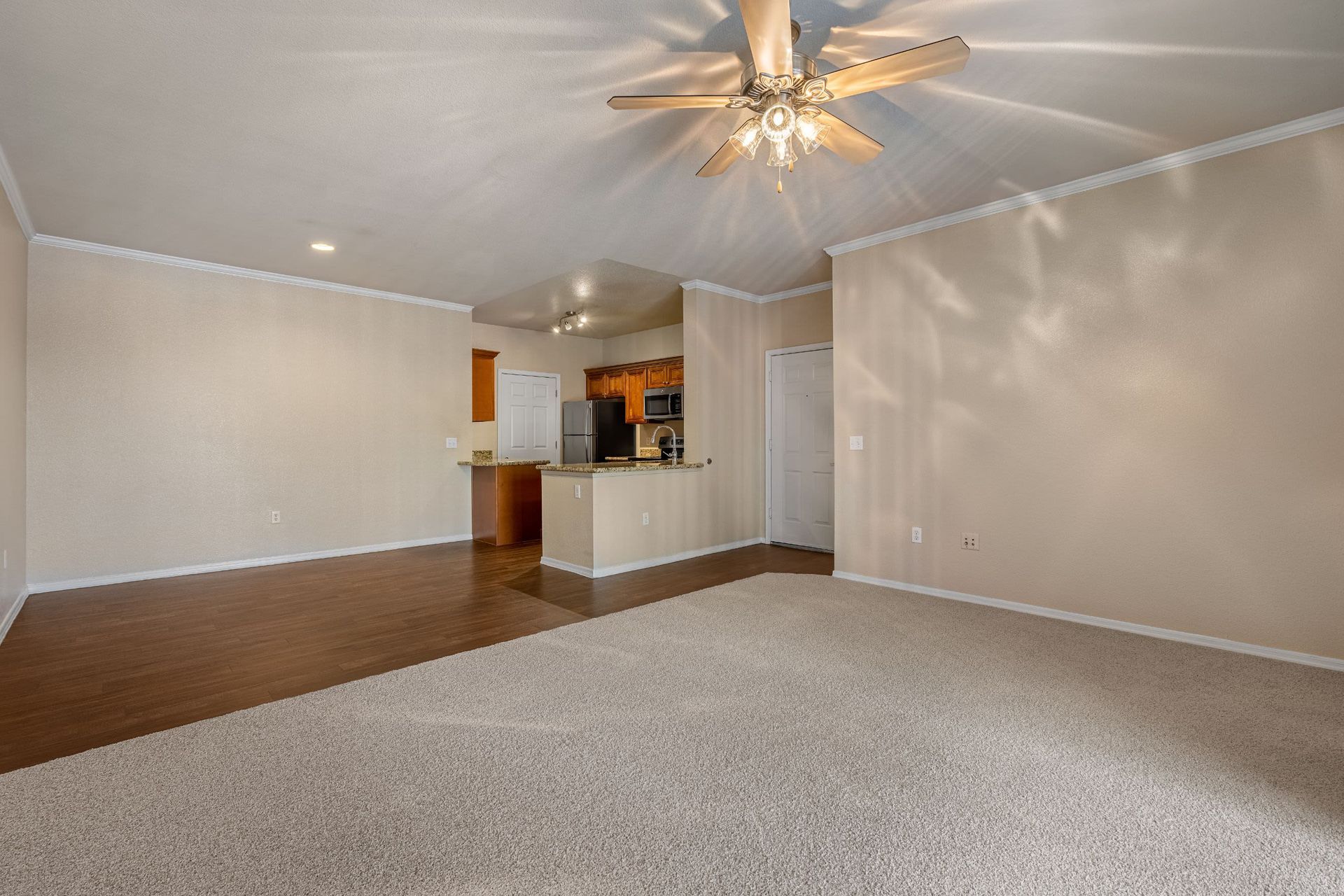 Spacious living room interior with beige walls, plush carpeting, and a dark hardwood floor section leading into a kitchen area with wooden cabinets and granite countertops. The room is illuminated by natural light and a ceiling fan with lights at Azure Creek in North Phoenix, AZ.