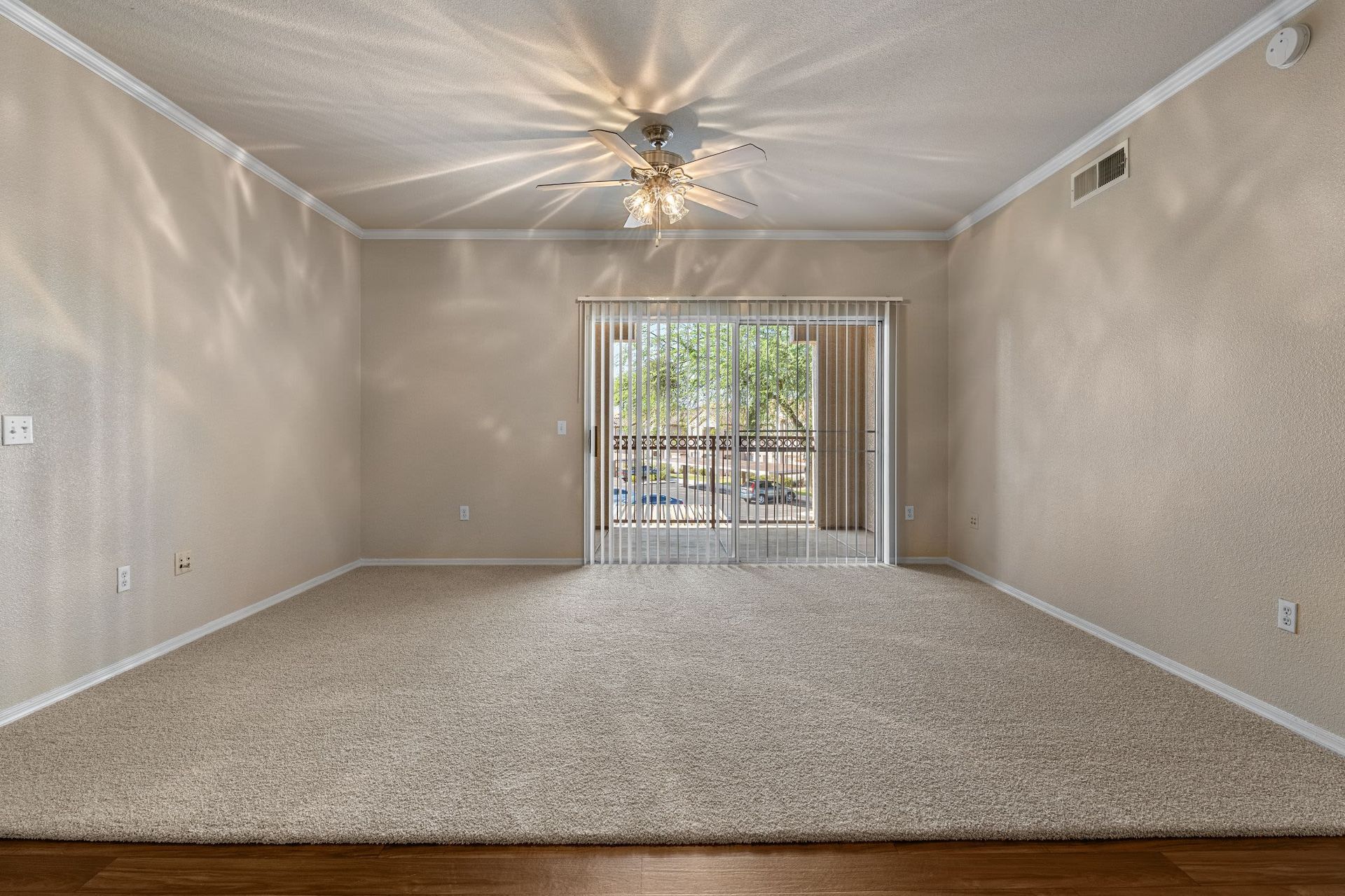 Empty living room with beige walls and carpet flooring, featuring a ceiling fan with lights casting a pattern on the ceiling, and large glass sliding doors with vertical blinds leading to an outside area at Azure Creek in North Phoenix, AZ.
