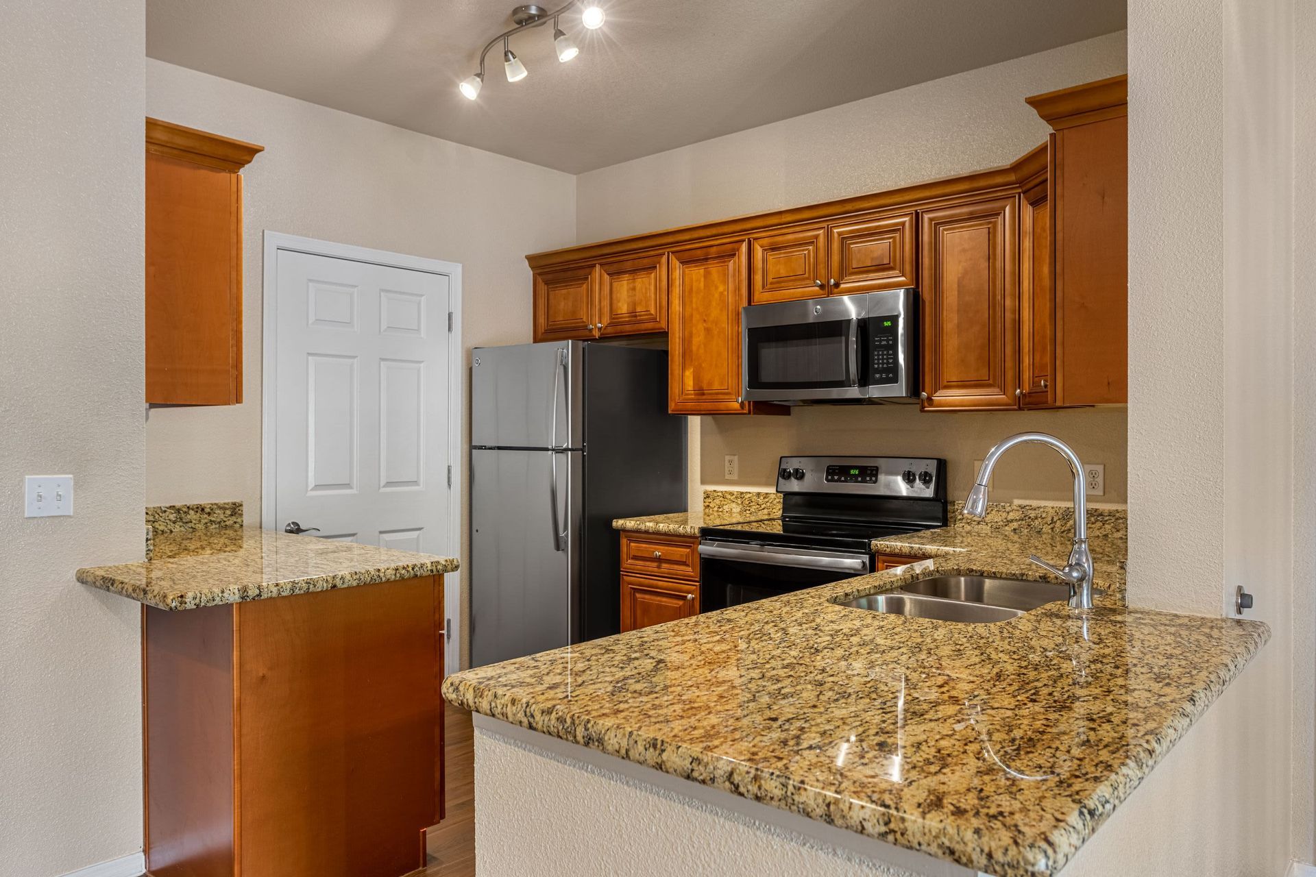 Modern kitchen interior featuring cherry wood cabinets, granite countertops, and stainless-steel appliances including a refrigerator, microwave, and range. A white paneled door leads to another room, and the kitchen is lit by a track lighting fixture on the ceiling at Azure Creek in North Phoenix, AZ.