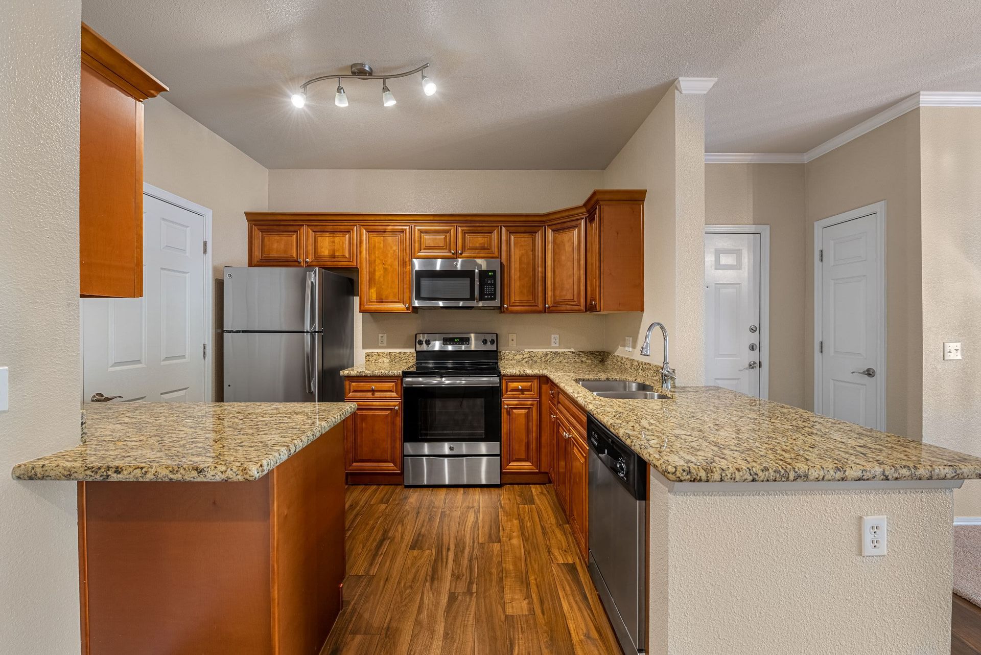 Modern kitchen interior with wooden cabinets and granite countertops, featuring stainless steel appliances, including a refrigerator, dishwasher, microwave, and oven. The room has hardwood flooring and is illuminated by ceiling spotlights. There are no people visible in the image at Azure Creek in North Phoenix, AZ.