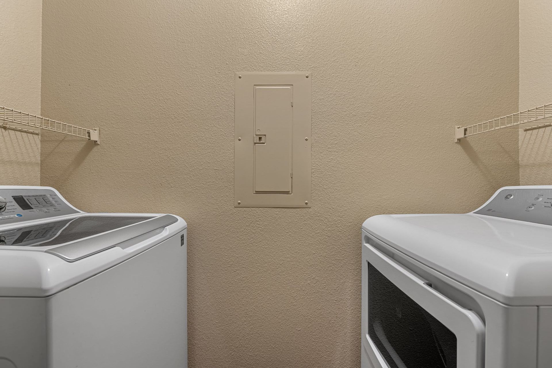 A laundry room with a beige wall featuring a central electrical panel flanked by a white top-load washing machine on the left and a matching white front-load dryer on the right. Wire shelving is installed above each appliance at Azure Creek in North Phoenix, AZ.