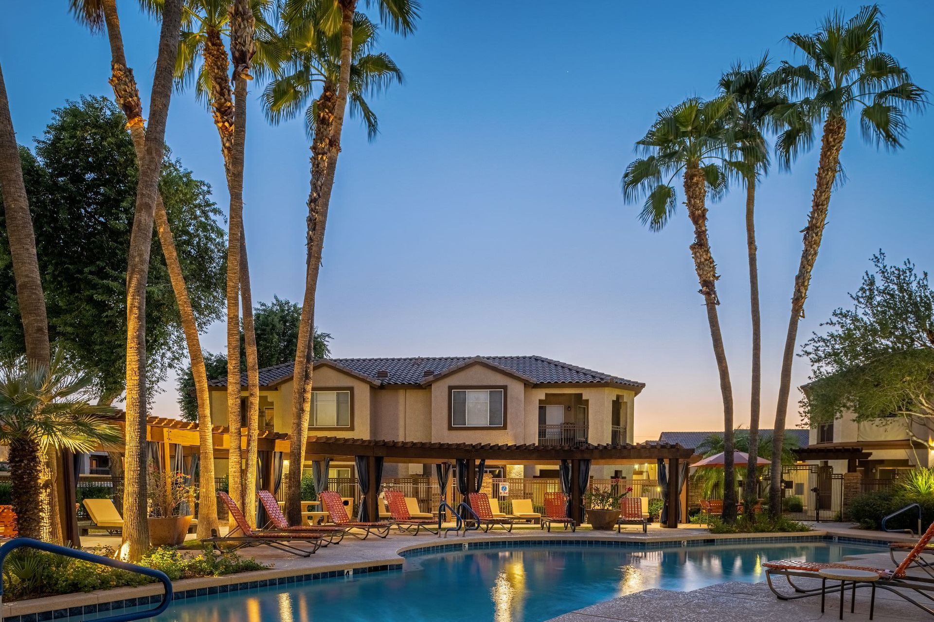 Twilight setting at a luxurious residential pool area surrounded by tall palm trees, with a two-story house in the background, poolside loungers, and a calm blue swimming pool reflecting the serene sky at Azure Creek in North Phoenix, AZ.