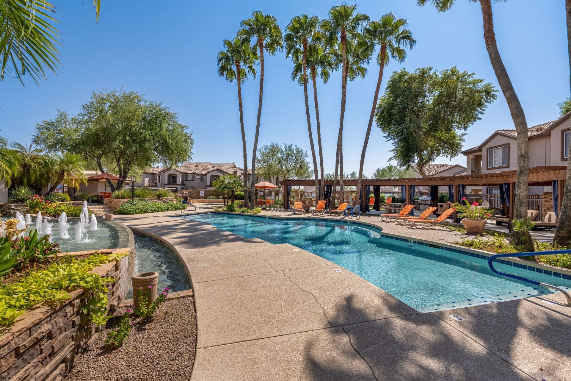 Resort-style outdoor swimming pool surrounded by palm trees, sun loungers, and a fountain, set against a backdrop of residential buildings with clear blue skies overhead at Azure Creek in North Phoenix, AZ.