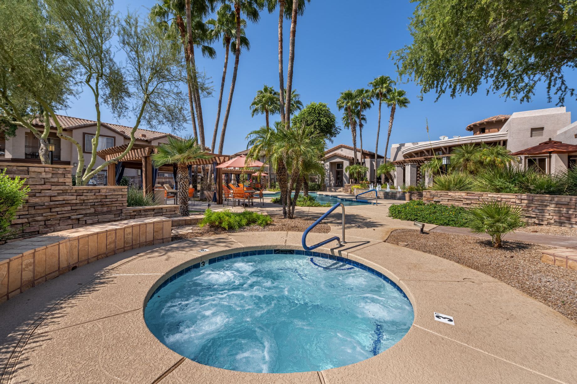 Relaxing resort-style outdoor area with a sparkling jacuzzi in the foreground, surrounded by a sun-drenched patio with loungers and tall palm trees. In the background, comfortable seating areas and elegant residential structures under a clear blue sky at Azure Creek in North Phoenix, AZ.