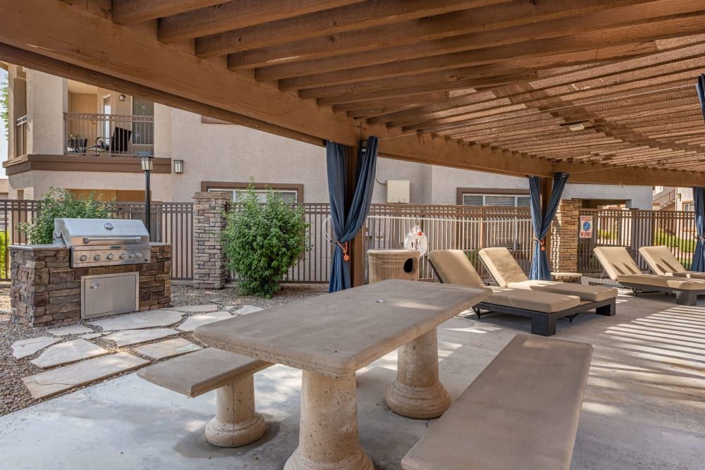 An outdoor covered patio featuring a built-in barbecue grill set against a stone wall, a long concrete table, and multiple sun loungers. The area is shaded by a wooden pergola and complemented by blue curtains. There's a fenced pool area visible in the background at Azure Creek in North Phoenix, AZ.