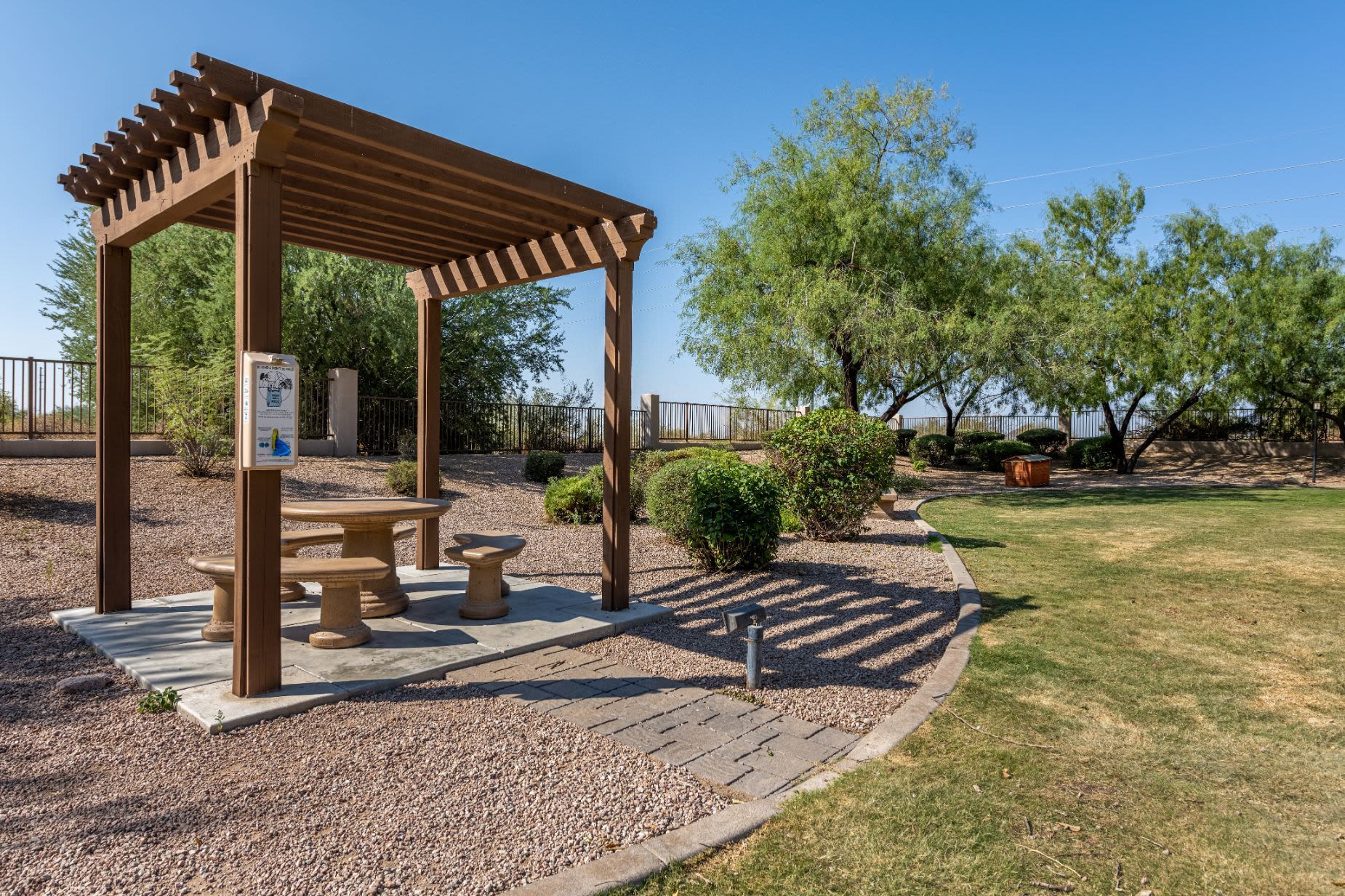 An outdoor rest area with a wooden pergola providing shade over a circular concrete table and two bench seats. The environment is a well-maintained park with gravel, green grass, a curved path with stone pavers, and mature trees in the background. A clear blue sky suggests a sunny day at Azure Creek in North Phoenix, AZ.