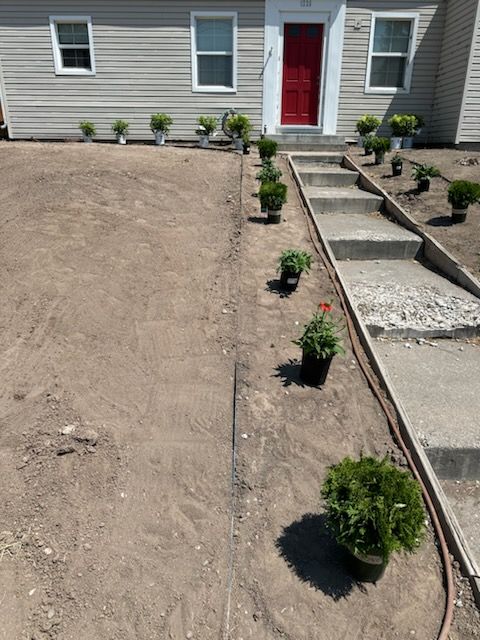 A view of a house exterior with a red front door, featuring newly planted shrubs along the walkway in a yard under repair.