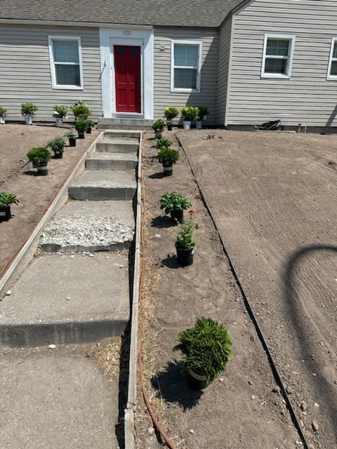 A front yard showing a concrete walkway lined with small potted plants and dirt, leading to a house with a red door.