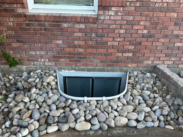 A semi-circular grey window well with a black cover, surrounded by decorative river stones against a brick house wall.
