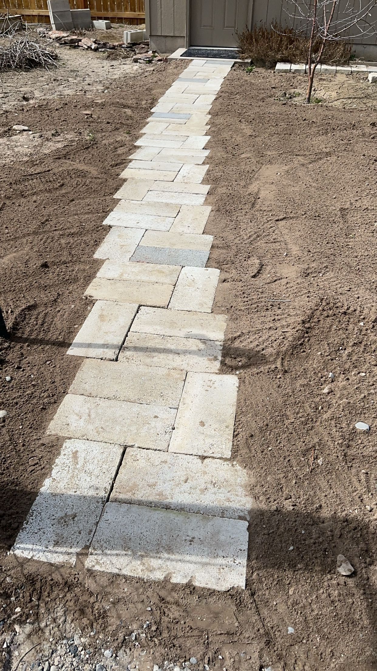 A stone pathway made of light-colored, rectangular pavers leading through a bare dirt yard toward a building entrance.