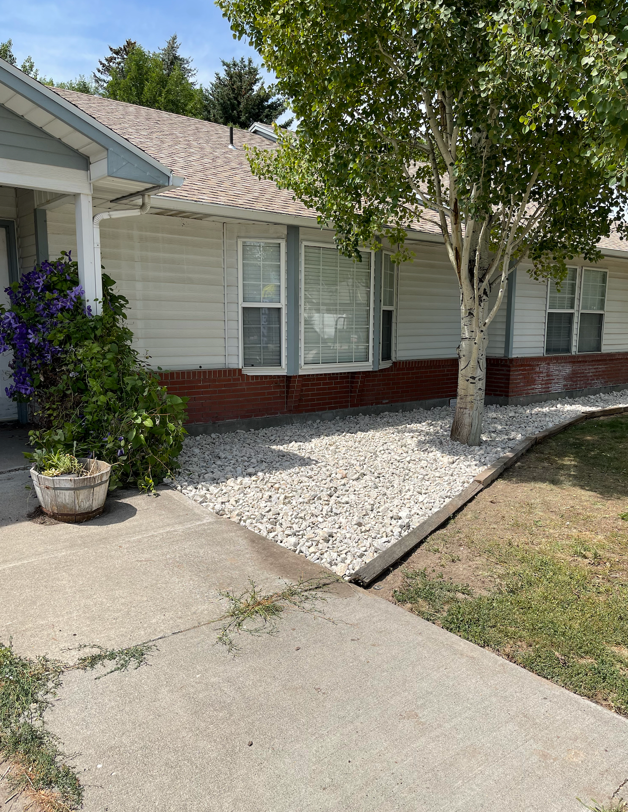 A one-story house with white horizontal siding and a red brick base, featuring a stone-filled yard and a large tree.
