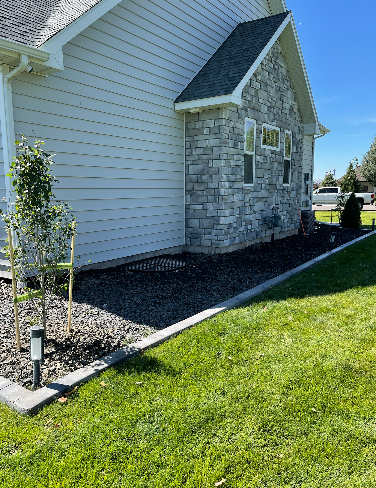 Side view of a house with light horizontal siding, a stone facade corner, dark gravel landscaping, and a green lawn.