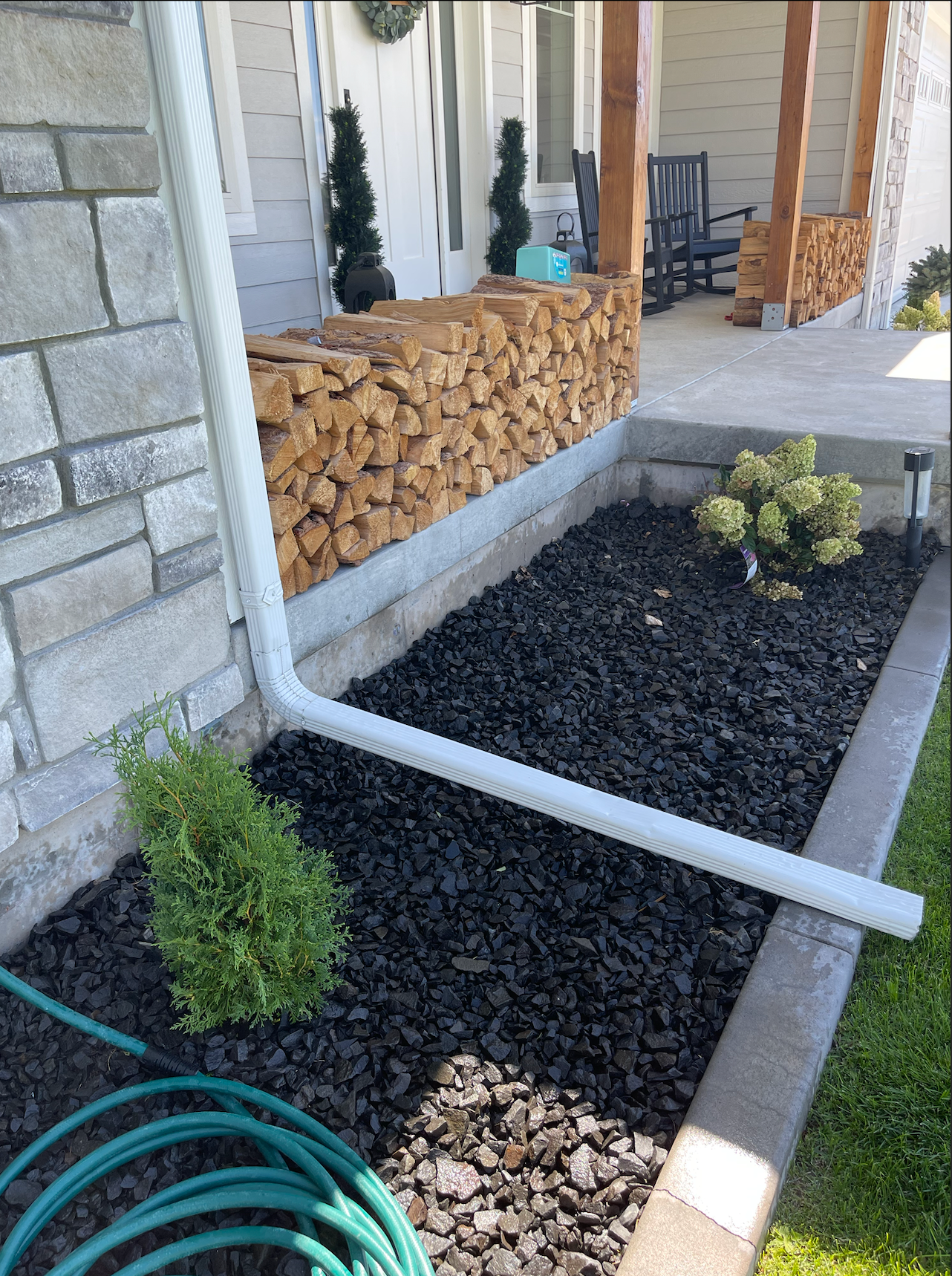 A white downspout extension lies across a dark mulch garden bed in front of a stone porch stacked with firewood.